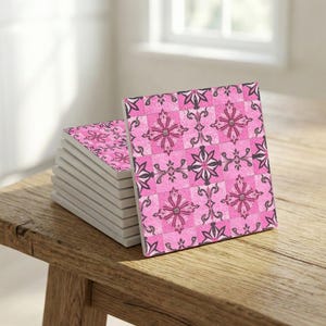 Puede incluir: Una pila de posavasos cuadrados con un estampado floral rosa. Los posavasos presentan un diseño repetitivo de flores rosas y detalles en gris oscuro sobre un fondo a cuadros. Los posavasos están apilados sobre una superficie de madera.