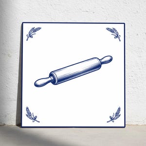 Puede incluir: Azulejo blanco cuadrado con borde azul e ilustración azul de un rodillo. Las esquinas presentan ramitas azules. El rodillo está centrado y tiene un estilo vintage detallado.