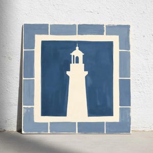 Puede incluir: Arte mural cuadrado con una silueta de faro blanco sobre un fondo azul oscuro. La obra de arte está enmarcada con un borde azul claro y un borde interior color crema, creando un tema náutico. La pieza se exhibe contra una pared blanca.