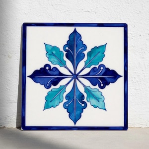 Op de afbeelding: Vierkante keramische tegel met een witte achtergrond en een symmetrisch bloemmotief in blauw- en groentinten. Het ontwerp is omlijst door een donkerblauwe rand. De tegel is waarschijnlijk voor decoratief gebruik.