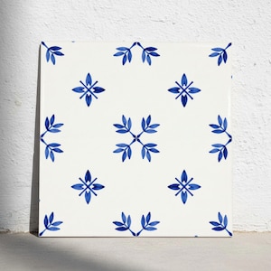 Puede incluir: Un azulejo de cerámica cuadrado con fondo blanco y un patrón repetitivo de motivos de hojas pintadas en azul. El diseño consiste en grupos simétricos de cuatro hojas dispuestas en forma de diamante, creando una cuadrícula visual uniforme.