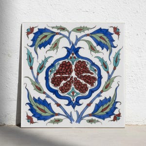 Könnte beinhalten: Eine quadratische Keramikfliese mit einer zentralen Granatapfel-Illustration in Dunkelrot, umgeben von komplexen blauen und grünen Blumenmotiven und geschwungenen Blättern auf weißem Grund. Das Design ist symmetrisch mit detaillierten Linien.