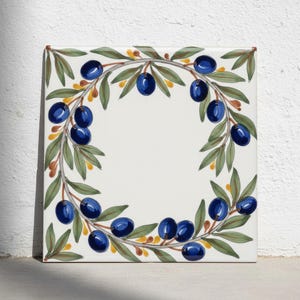 Puede incluir: Un azulejo de cerámica cuadrado con un diseño de corona pintado a mano de aceitunas azul oscuro y hojas verdes sobre un fondo blanco. Las ramas de olivo están dispuestas en un patrón circular, acentuadas con pequeños detalles en amarillo y marrón.