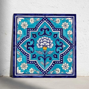 Puede incluir: Azulejo cerámico cuadrado con diseño floral. El azulejo presenta una flor central en blanco y azul, rodeada de un fondo turquesa y un borde azul oscuro. Pequeñas flores blancas y puntos decoran los bordes. El azulejo mide aproximadamente 30 cm cuadrados.