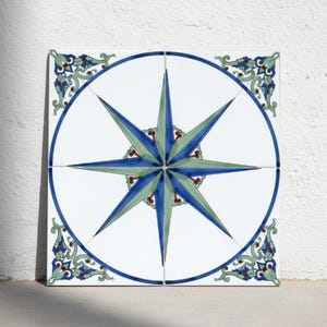 Puede incluir: Un mural de azulejos decorativos cuadrado compuesto por cuatro piezas individuales que presenta un diseño central de rosa de los vientos en tonos azules y verdes. El patrón está encerrado en un borde circular azul con adornos florales en las esquinas sobre blanco.