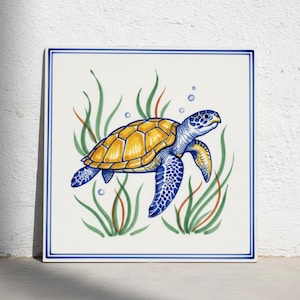 Puede incluir: Un azulejo de cerámica cuadrado con una ilustración pintada a mano de una tortuga marina con caparazón amarillo y aletas con patrones azules. La tortuga está rodeada de algas verdes y pequeñas burbujas azules, enmarcada por un borde azul.