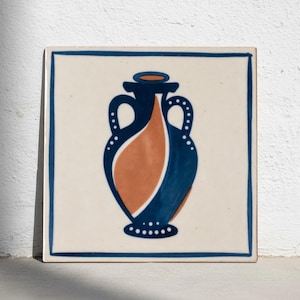 Puede incluir: Azulejo cerámico cuadrado con un diseño de ánfora estilizada. El jarrón es azul marino y naranja, con detalles de puntos blancos. El azulejo tiene un fondo color crema y un borde azul marino.