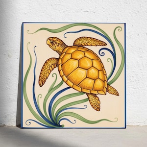Puede incluir: Un azulejo de cerámica cuadrado que presenta una tortuga marina amarilla pintada a mano nadando entre algas verdes y azules estilizadas. El diseño está sobre un fondo de color crema y enmarcado por un borde fino de color azul oscuro.