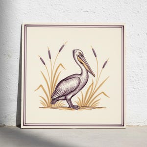 Op de afbeelding: Een vierkante decoratieve tegel met een gedetailleerde illustratie van een pelikaan die tussen hoog riet staat. De vogel is weergegeven in paarse en crèmetinten, met een gele snavel. Het ontwerp is omlijst door een dunne paarse rand op een crèmekleurige achtergrond.