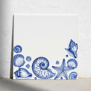 Op de afbeelding: Een vierkante witte keramische tegel met een decoratieve rand van blauwe illustraties van diverse schelpen en een zeester. Het ontwerp bevat een grote centrale nautilusschelp, een mantelschelp en een hoornschelp langs de onderrand.