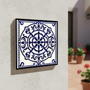May include: Square ceramic tile with a black frame, featuring a symmetrical blue and white floral design. The tile is mounted on a white wall, showcasing a decorative element with intricate patterns and a classic aesthetic.