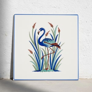 Puede incluir: Un azulejo de cerámica cuadrado con una ilustración central de un flamenco azul de pie sobre una pata entre juncos altos verdes y azules con espadañas de color marrón rojizo. El azulejo tiene un fondo blanco y un borde azul fino.