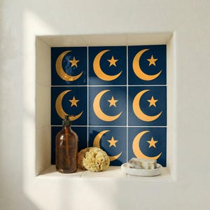 Può includere: Una nicchia del bagno presenta piastrelle blu navy con disegni dorati a forma di luna crescente e stella. Un dosatore di sapone marrone, una spugna naturale e un portasapone con sapone sono sullo scaffale. Le piastrelle creano uno sfondo decorativo.