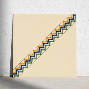Puede incluir: Un azulejo cuadrado de cerámica con una base de color crema. Una banda diagonal recorre desde la esquina inferior izquierda hasta la superior derecha, con un patrón de zigzag repetido en franjas alternas amarillas, azules y verdes, bordeadas por finas líneas negras.