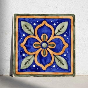 May include: Square ceramic tile with a floral design. The tile features a blue background with an orange flower in the center, surrounded by green leaves. The border is green and orange. White dots are scattered around the flower.