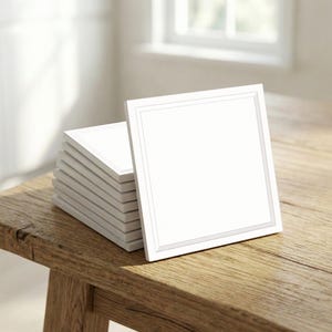 Puede incluir: Una pila de posavasos cuadrados blancos con un fino borde gris, colocados sobre una superficie de madera rústica. Los posavasos están apilados, con uno inclinado contra la pila. La escena está iluminada por luz natural.