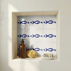Puede incluir: Un nicho de ducha empotrado con azulejos blancos con un diseño de pez azul. Un dispensador de jabón marrón, una esponja natural y un jabonera con jabón están en el estante. El diseño tiene un tema náutico.