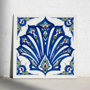 May include: A square ceramic tile featuring a symmetrical floral pattern in shades of cobalt blue, turquoise, and sage green on a white background. The design includes intricate petal-like shapes and small red accents in the corners and center.