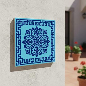 Puede incluir: Azulejo cuadrado azul claro con un diseño floral azul oscuro en el centro y un borde geométrico. El azulejo está montado en una pared blanca, con una vista de un patio y plantas en macetas en el fondo.