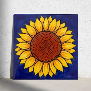 May include: Square ceramic tile with a sunflower design. The sunflower has yellow petals with orange shading, a brown centre, and is set against a deep blue background. The artwork is a decorative piece.