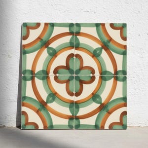 Könnte beinhalten: Eine quadratische Dekofliese mit einem handgemalten geometrischen Muster in Salbeigrün und Terrakotta-Orange auf cremeweißem Grund. Das Design besteht aus ineinandergreifenden kreisförmigen Bögen und einem zentralen vierblättrigen Blumenmotiv.