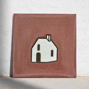Puede incluir: Azulejo cerámico cuadrado con un diseño de casa blanca sobre un fondo rosa empolvado. La casa tiene un contorno y detalles negros, incluyendo una puerta, ventanas y una chimenea. La baldosa es probablemente para uso decorativo.
