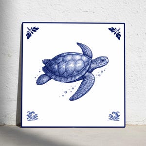 Op de afbeelding: Vierkante keramische tegel met een gedetailleerde blauwe illustratie van een zwemmende zeeschildpad. De tegel heeft een witte achtergrond, een blauwe rand en decoratieve hoekaccenten. De schildpad is het centrale punt.