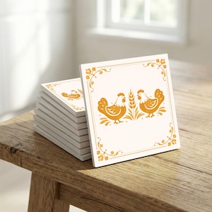 Könnte beinhalten: Ein Stapel quadratischer Keramikuntersetzer. Jeder Untersetzer hat einen cremefarbenen Hintergrund mit einem orangefarbenen Design aus zwei Hühnern und Weizen. Das Design wird von einem orangefarbenen Blumenrand umrahmt. Die Untersetzer liegen auf einer Holzoberfläche.