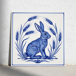 May include: A square ceramic tile featuring a blue and white illustration of a rabbit sitting in the centre, surrounded by a circular arrangement of wheat stalks. The design is framed by a thin blue border on a white background.