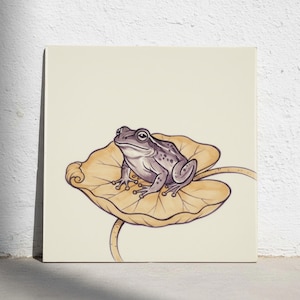 Puede incluir: Una impresión artística cuadrada que presenta una ilustración detallada de una rana de color morado claro con manchas oscuras sentada sobre un nenúfar grande de color amarillo dorado. La rana está representada de perfil, mirando hacia la izquierda.