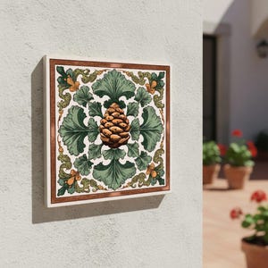 Può includere: Piastrella decorativa quadrata con un'illustrazione dettagliata di una pigna circondata da foglie verdi e accenti floreali. Il design è incorniciato da un bordo marrone e posto su uno sfondo bianco. La piastrella è montata su un muro bianco.