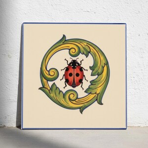 May include: A square ceramic tile with a cream-colored background and a thin blue border. In the center, a red ladybug with black spots is framed by a circular, ornate scrollwork design featuring vibrant shades of yellow and green leaves.