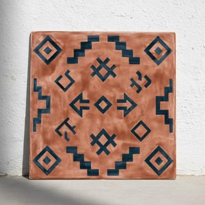 Puede incluir: Azulejo cuadrado de terracota con un patrón geométrico en azul oscuro. El diseño presenta diamantes, flechas y formas escalonadas. El azulejo está apoyado contra una pared blanca.