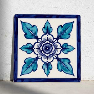Puede incluir: Azulejo cerámico cuadrado con diseño floral. El azulejo presenta un fondo blanco con una flor central y hojas circundantes en tonos azules. Un borde azul oscuro enmarca el diseño.