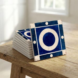 Puede incluir: Una taza de cerámica blanca con asa negra y el texto "But First, Coffee" en negrita negra. La taza se encuentra sobre un fondo borroso, sugiriendo un ambiente acogedor.