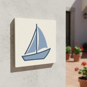 Puede incluir: Una pieza de arte mural cuadrada, de color blanquecino, con una ilustración de un velero azul. El velero tiene un casco y una vela azules, con un contorno azul oscuro. La obra de arte está montada en una pared clara y texturizada.