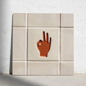May include: Square ceramic tile with a cream-colored background and a raised, brown hand gesture in the center. The hand is making the "okay" sign. The tile is surrounded by a grid of smaller cream-colored tiles.