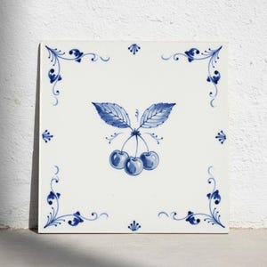 May include: A square ceramic tile with a white background and a hand-painted blue design. The center features three cherries with two leaves on stems. The corners and edges are decorated with delicate blue scrollwork and floral motifs.