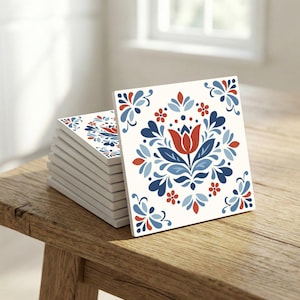 Puede incluir: Una pila de baldosas cerámicas cuadradas con un diseño floral. El diseño presenta una flor central roja en forma de tulipán rodeada de hojas y flores estilizadas azules y rojas sobre un fondo blanco. Las baldosas están apiladas sobre una superficie de madera.