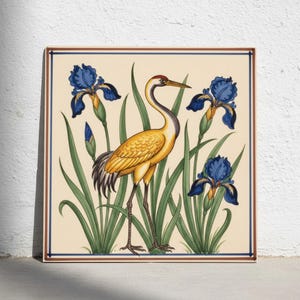 May include: A square ceramic tile featuring a central yellow crane standing among green leaves and three blue iris flowers. The design is set against a cream background and framed by a thin blue and brown border, creating a decorative wall art piece.