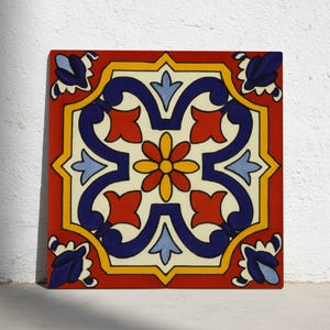 Può includere: Piastrella quadrata in ceramica con un vivace motivo floreale. La piastrella presenta un bordo rosso, un disegno centrale giallo e crema e accenti floreali blu e rossi. Il fiore centrale è giallo e rosso.