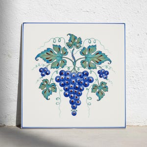 May include: A square ceramic tile featuring a hand-painted illustration of a cluster of dark blue grapes with light blue highlights. The grapes are surrounded by teal and green vine leaves and curling tendrils, all set against a white background.