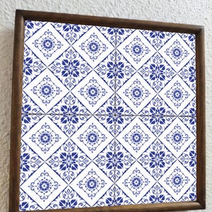 Puede incluir: Una bandeja cuadrada de madera con un diseño de azulejos decorativos. Los azulejos tienen un fondo blanco con un patrón floral azul repetido. El marco de la bandeja es de color marrón oscuro.