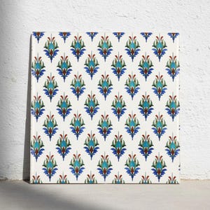 Puede incluir: Un azulejo de cerámica cuadrado con un patrón repetitivo de motivos florales estilizados en tonos turquesa, azul profundo y rojo oscuro sobre un fondo blanco sólido. El diseño consta de formas simétricas similares a hojas puntiagudas.