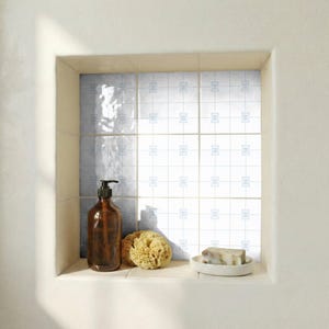 Puede incluir: Un nicho de baño con un dispensador de jabón de vidrio marrón, una esponja natural y una pastilla de jabón en un plato blanco. El fondo presenta azulejos blancos con un patrón floral azul. El nicho está enmarcado por una pared blanca.