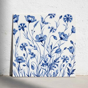 May include: A square ceramic tile featuring a white background with a repeating pattern of blue botanical illustrations. The design includes various wildflowers, cornflowers, and delicate stems with leaves, rendered in shades of blue.