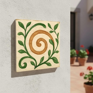 Puede incluir: Azulejo cuadrado de color crema con un diseño de espiral marrón en el centro, rodeado de vides de hojas verdes. El azulejo está montado en una pared blanca, mostrando una estética decorativa inspirada en la naturaleza.
