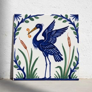 May include: A square ceramic tile featuring a hand-painted dark blue heron with wings partially spread. The bird holds a small orange swirl in its beak. The scene is framed by blue leafy vines, green reeds, and orange cattails on a white background.