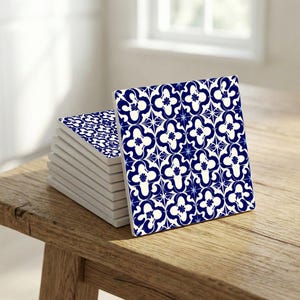 Puede incluir: Una pila de posavasos cuadrados de cerámica con un patrón floral azul y blanco. Los posavasos están sobre una mesa de madera. El patrón presenta flores estilizadas y formas de estrella. Miden aproximadamente 10 cm x 10 cm.
