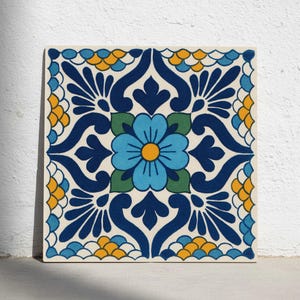May include: Square ceramic tile with a floral design. The tile features a central blue flower with a yellow centre, surrounded by green leaves and intricate blue and yellow patterns on a cream background.